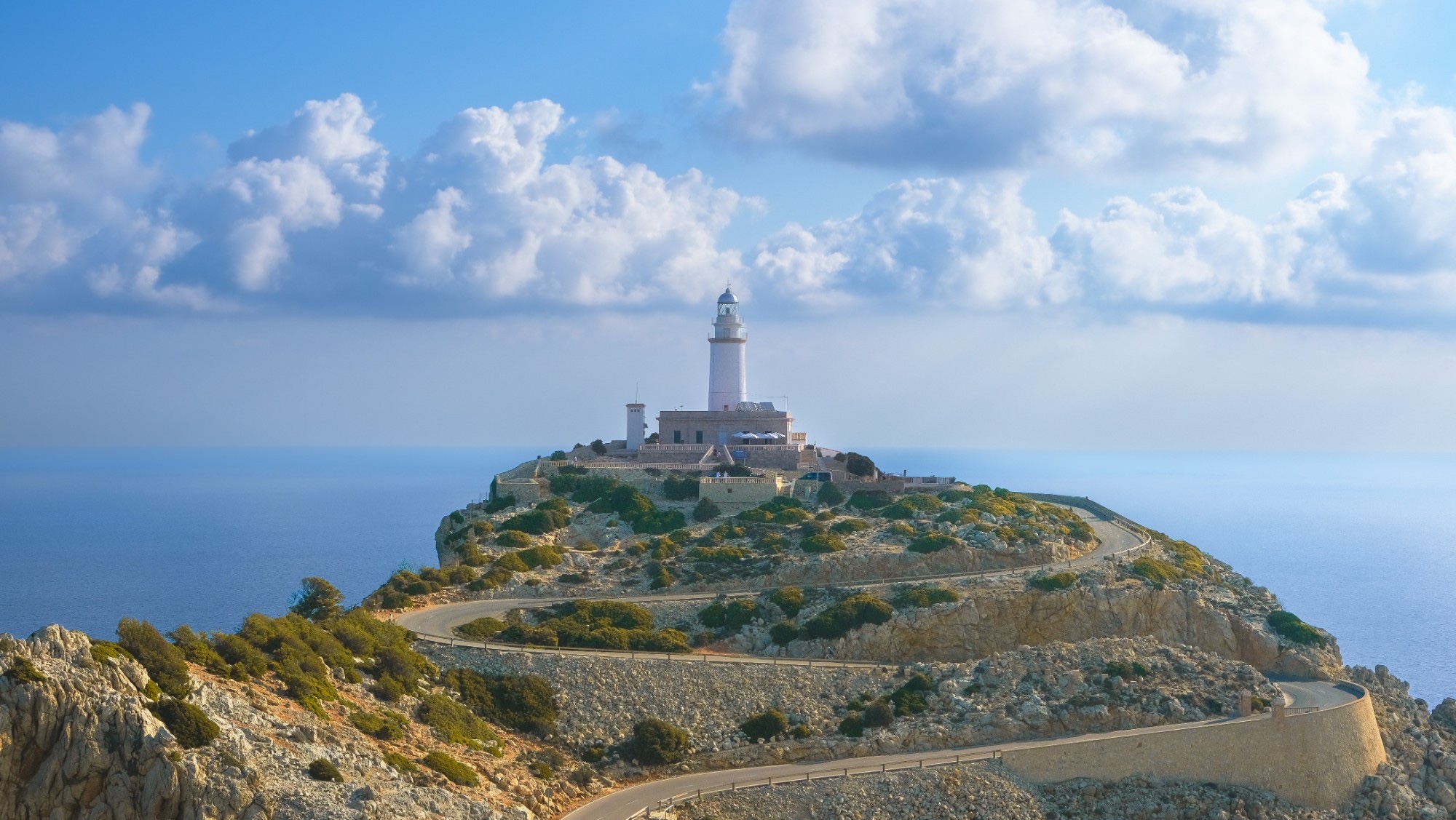A white lighthouse on a rocky headland above the sea, under big summer clouds.